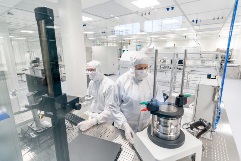 Industriefotografie: Blick in einen Reinraum zur Fertigung von elektronischen Leuchtelementen bei einem Hersteller von Leuchtmitteln. Zwei Mitarbeiter in weissem Overalls mit Haube und Gesichtsmasken prüfen Bauteile.