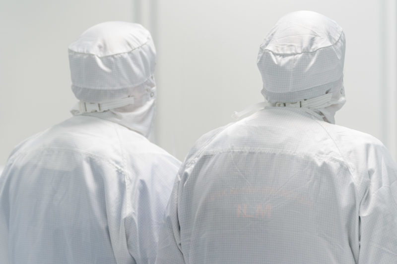 Industrial photography:  Two employees of a clean room photographed from behind. Through their white overalls and hoods the whole picture with the white background seems a bit unreal.