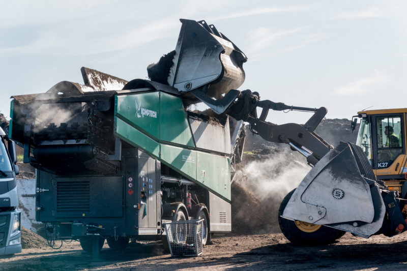 Reportage photography: Backlight photo of a screening machine for compost waste, which is currently being filled with material by a shovel loader.