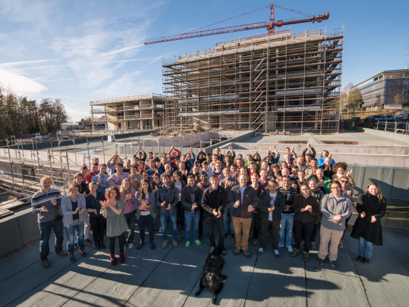 Group photo: The future users of a new building celebrate the shell construction of the building. They applaud while in the background the building with construction crane and scaffolding can be seen. In the foreground is the dog, which is allowed to move into the new office during the working hours of its owner.