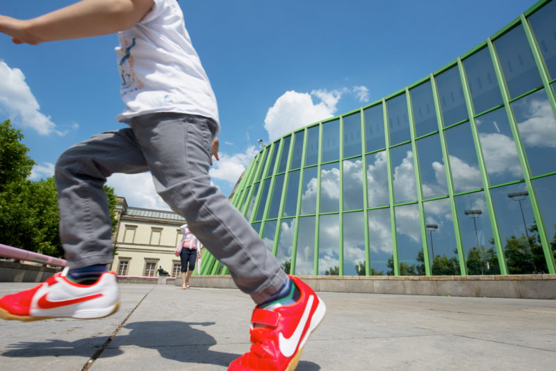 Ein Junge mit roten Turnschuhen springt über den Vorplatz der Staatsgalerie Stuttgart. In der geschwungenen Glasfront des Foyers spiegeln sich die Wolken vor dem blauen Himmel. Im Hintergrund eine Passantin und ganz klein sichtbar die Plastik Draped Reclining Woman von Henry Moore.