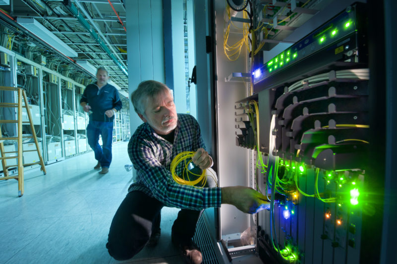 Industrial photography: Press photo on the subject of IT, data and communication: In a control room for data lines, an employee connects a fiber optic cable to a switch. The LEDs on the devices bathe the room in green light. A colleague walks in the background along many connecting cables on the wall and ceiling.