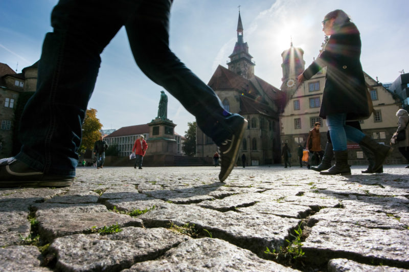 Stadtportrait Stuttgart: Auf dem Schillerplatz in Stuttgart laufen Passanten mit großen Schritten über den sonnendurchfluteten Platz. Im Hintergrund die Silhouette der Stiftskirche.