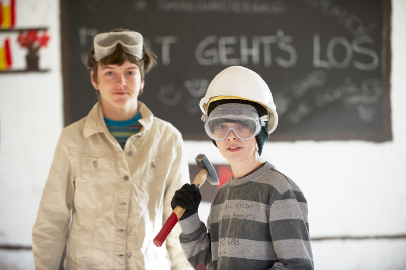 Editorial photography: Two students in an educational project helping to renovate an old house. The boy in the foreground wears a construction helmet and a heavy hammer. They both wear safety goggles.