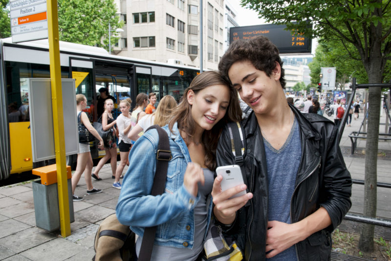 Lifestyle-Fotografie: Eine Schülerin und ein Schüler schauen an einer Bushaltestelle in ein Smartphone.