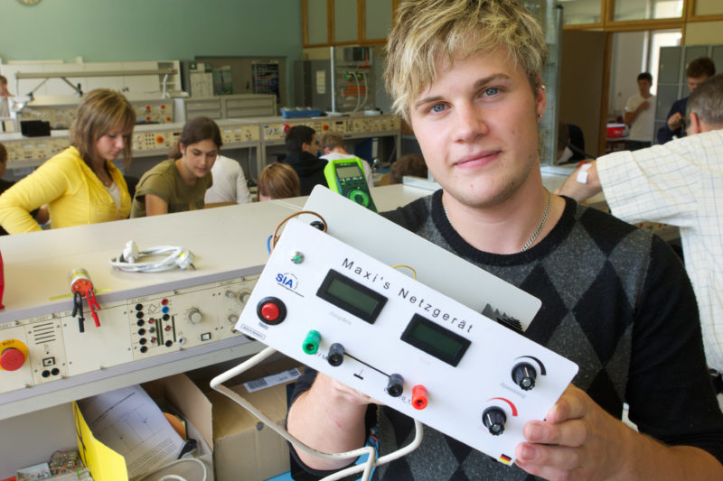 Reportagefotografie: An einer weiterführenden Schule arbeiten Schüler im Elektoniklabor. Einer hält sein selbstgebautes Netzgerät in die Kamera, das mit seinem Namen beschriftet ist.