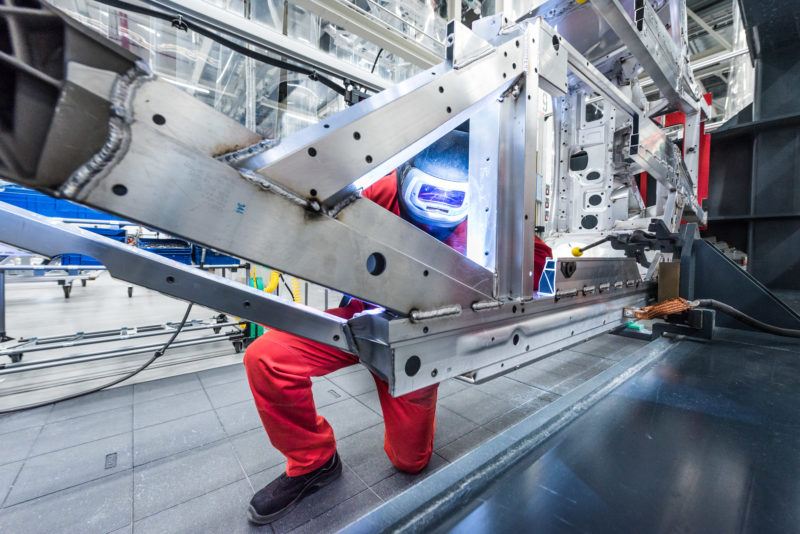 Industriefotografie: Ein Schweisser mit einem Schutzhelm arbeitet an einer Fahrzeugkarosserie. Man sieht den Lichtschein der blauen Schweissflamme im Helm und das glänzende Aluminium der Karosserie.