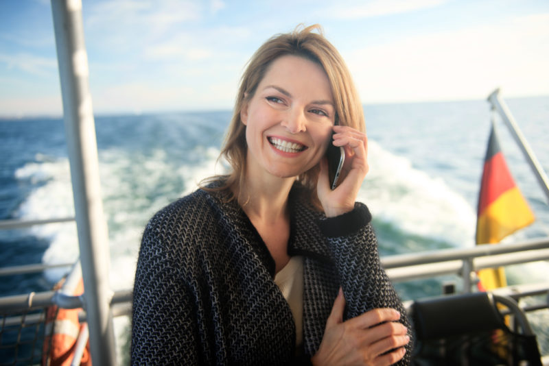 Industriefotografie: Eine Frau telefoniert mit ihrem Smartphone an Bord eines Schiffes. Im Hintergrund die deutsche Fahne am heck des Bootes.