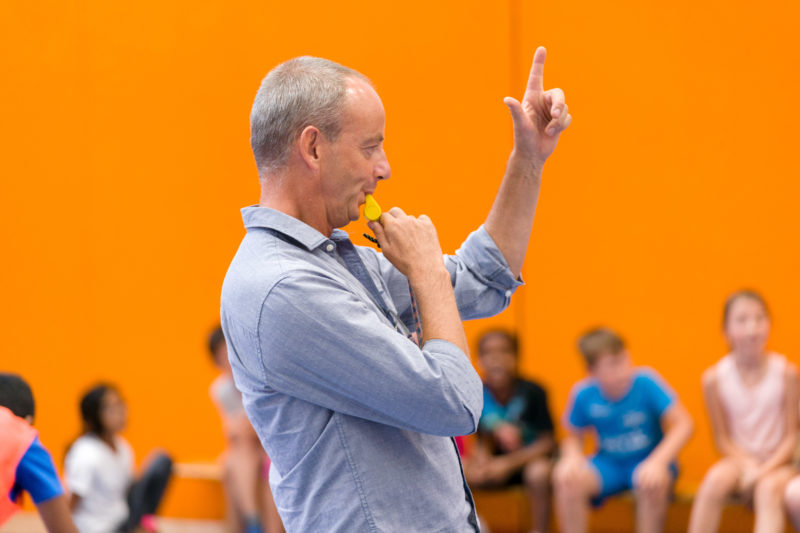 Reportagefotografie: Ein Lehrer bei der Arbeit: Reportagefotografie zum Thema Lernen und Bildung: Ein Sportlehrer benutzt in der Sporthalle seine Trillerpfeife und hebt seinen Zeigefinger, um die Aufmerksamkeit seiner Klasse zu bekommen.