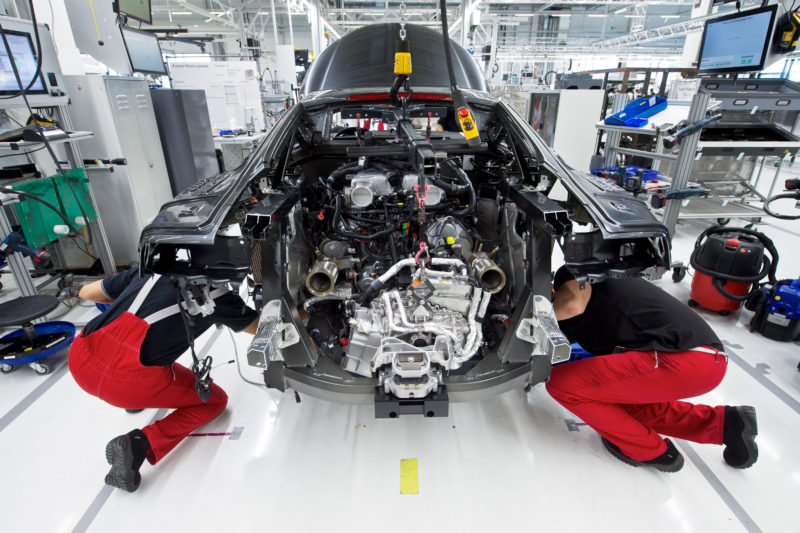 Industriefotografie: Zwei Mitarbeiter in einer Autommobilmanufaktur befestigen den Benzinmotor eines Sportwagens an seinem Platz im Heck des Fahrzeuges. Dazu müssen sie sich tief in das Fahrzeug hineinbeugen. Man sieht das komplizierte Aggregat  mit seinen vielen silbernen und schwarzen Bauteilen.