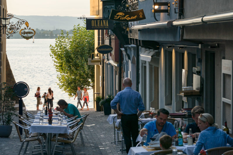 Stadtportrait: Zug am Zuger See. Menschen essen draußen vor den Restaurants, die an einer Gasse liegen, die zum See führt.