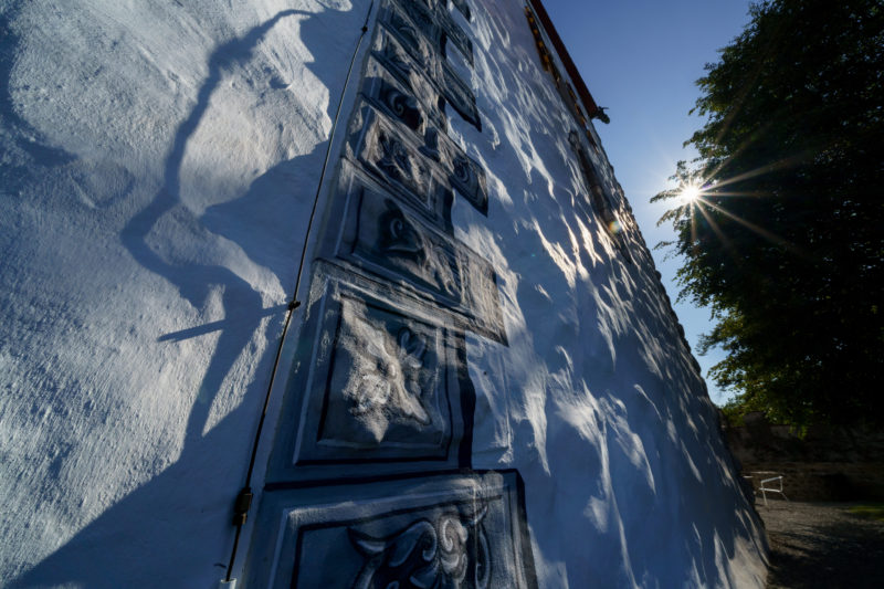 Stadtportrait: Zug am Zuger See. Schattenspiel auf den gemalten Steinen einer alten Gebäudemauer im historischen Stadtkern. Die Sonne scheint durch die Äste eines alten Baumes.