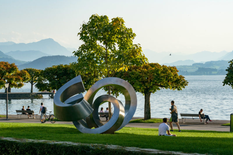 Portrait of the town: Train at Lake Zug. A sculpture made of shiny metal on the promenade on Lake Zug with strollers and people in their leisure time.