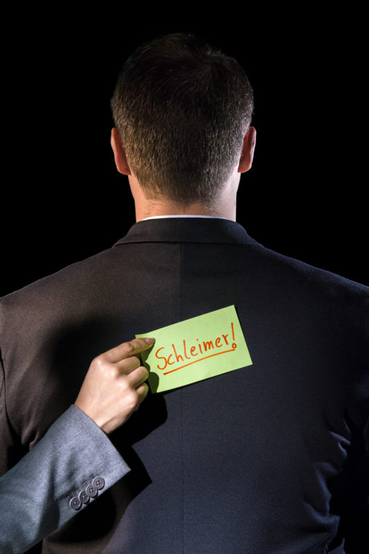 Studio photogaphy: Symbol photo on the subject of employee culture. A hand sticks a Post-It on the back of a man in a suit. It has the word suck-up written on it.