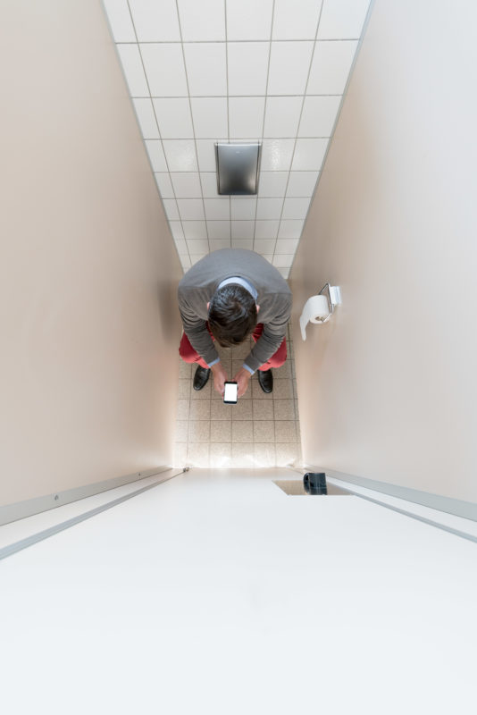 Studio photogaphy: A man sits in a toilet cubicle and looks into his smartphone. The picture was taken vertically from above.