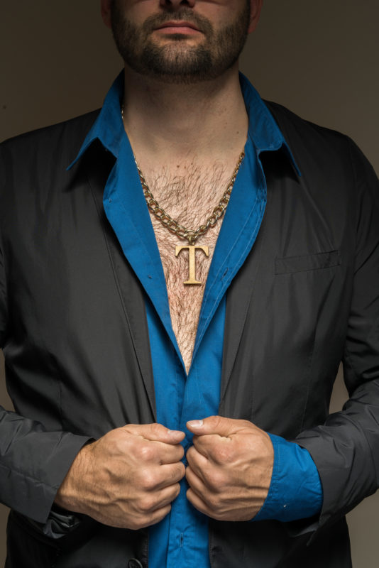 Studio photogaphy:  Symbol image for a gripping type. He has dark thick chest hair, wears an open shirt and a large amulet with a company logo around his neck.