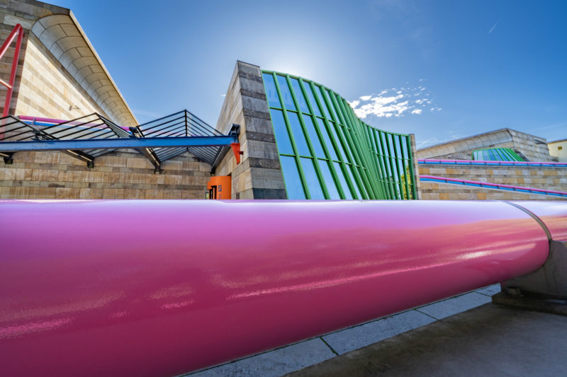 Stadtportrait Stuttgart: Die berühmte von James Stirling entworfene postmoderne Architektur der Eingangsfront der Neuen Staatsgalerie Stuttgart mit ihrer geschwungenen grün eingefassten Glasfront und der Metallbrüstung in rosa. Dahinter der blaue Himmel und die dazugehörigen Gebäudeteile aus Travertin und Sandstein.