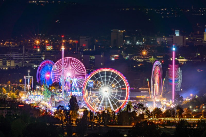 Stadtportrait Stuttgart: Eine Nachtaufnahme der beleuchteten Fahrgeschäfte beim Volksfest auf dem Cannstatter Wasen. Vor allem die Riesenräder zeichnen schöne Spuren in bunten Farben.