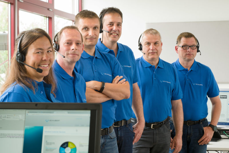 Group photo: Six employees of a company stand one behind the other in a row. All wear the blue T-shirt with the company name. The photo is taken with a light telephoto lens with tilt mechanism so that the oblique focus is on all faces.