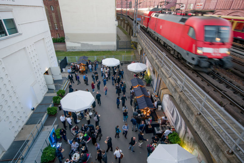 Reportagefotografie als Eventfotografie und Messefotografie: Bei einem Firmenevent im Umspannwerk Alexanderplatz in Berlin donnern Züge die angrenzende erhöht liegende Gleisanlage entlang. Unten versorgen die Teilnehmer im Hof der Eventlocation an kleinen Zelten mit Essen.