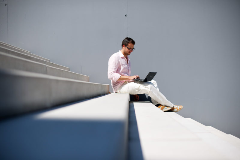 Lifestyle-fotografie: Ein Mann sitzt draußen auf einer Treppe in der Sonne und arbeitet mit seinem Notebook.