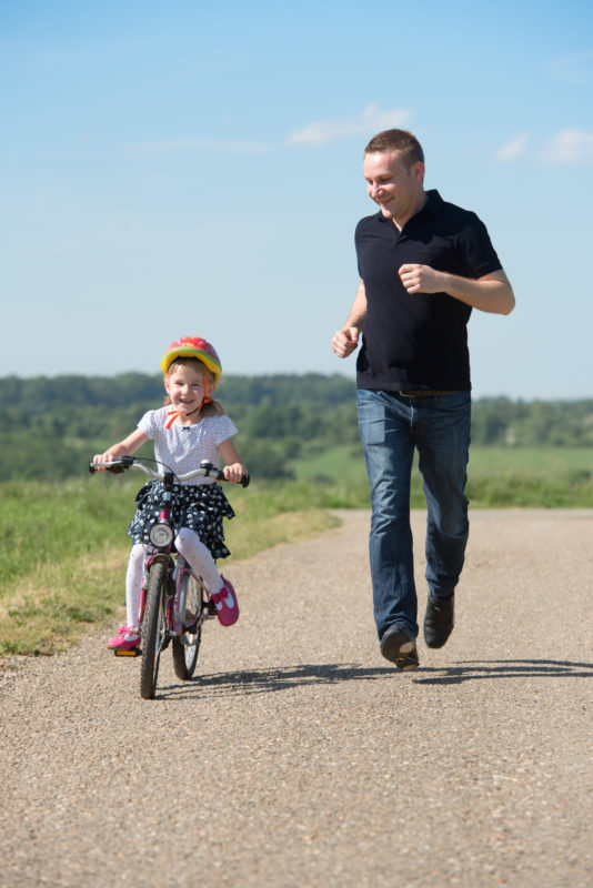 Industriefotografie: Reportagefotografie zu dem Thema Lernen und Bildung: Reportagefotografie: Ein Vater bringt seiner kleinen Tochter auf einem Feldweg in der Sonne das Fahrradfahren bei.