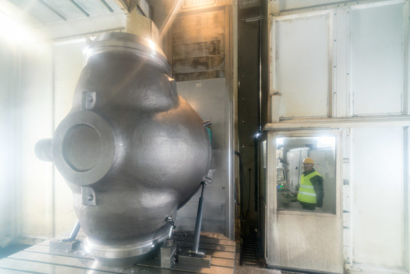 Industrial photography: A valve housing for a giant pipeline valve is machined in a closed milling machine. The machine operator with protective goggles, helmet and signal vest looks through a window into the plant.