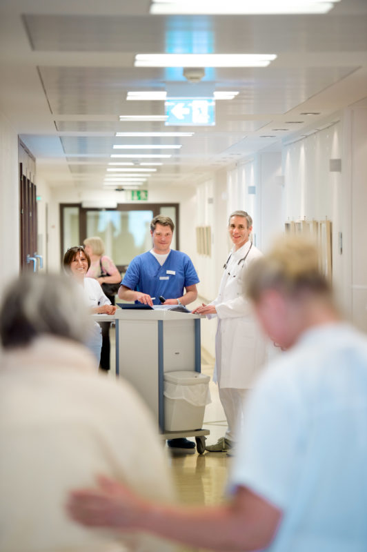 Medizinfotografie: Visite in einem Krankenhaus. Zwei Ärzte und eine Krankenschwester bei der Fallbesprechung auf dem Flur. Eine Patientin im Vordergrund wird von einer Krankenschwester geführt.