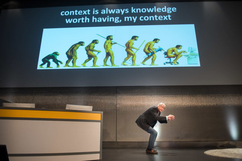 Editorial photography as event photography and fair photography: In a presentation to IT scientists, the speaker imitates a person on a computer. On the beamer picture you can see a drawing showing the development of humans from monkeys and their return to the bent monkey posture in front of the computer.