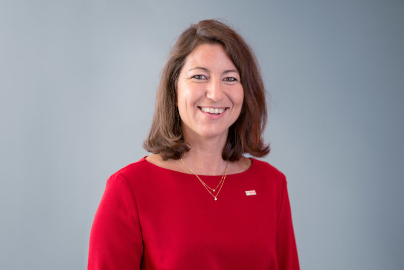 Managerportrait: A female manager in a red sweater against a grey background. She smiles into the camera and wears a small badge of her company and jewellery.