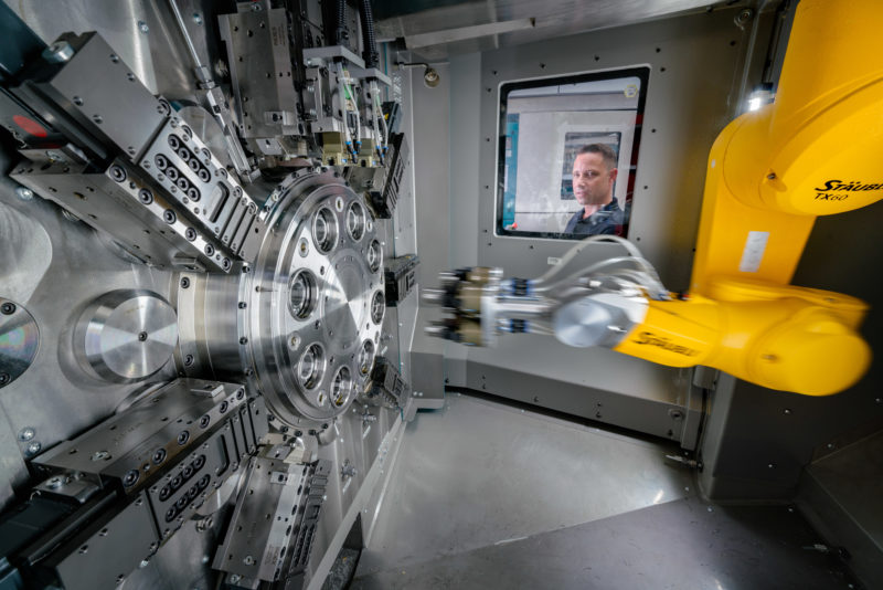 Industriefotografie: Ein gelber Roboterarm arbeitet in einer Werkzeugmaschine an einem großen Metallteil. Ein Mitarbeiter schaut durch die Glasscheibe in das Maschineninnere.