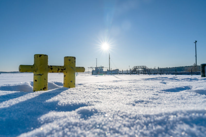 Landschaftsfotografie: Winter in Finnland: Ein Bootssteg führt zu einem kleinen Fischerboot, das im Wintereis eingefroren ist.  Die tiefstehende Sonne strahlt am blauen Himmel.