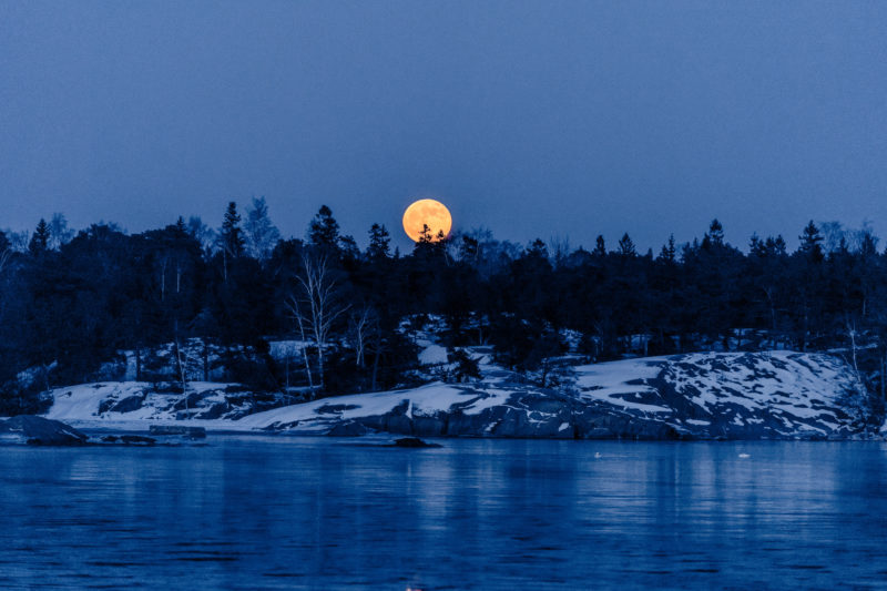 Landschaftsfotografie: Winter in Finnland: Der aufgehende rote Mond über den typischen schneebedeckten Felsen der Ostseeküste.