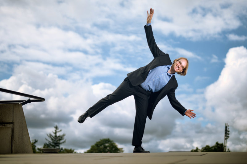 Mitarbeiterfotografie: Ein Wissenschaftler, der auch über künstliches Gleichgewicht forscht, beim Portrait draußen. Er balanciert auf einem Bein, während im Hintergrund Wolken vorbeiziehen.