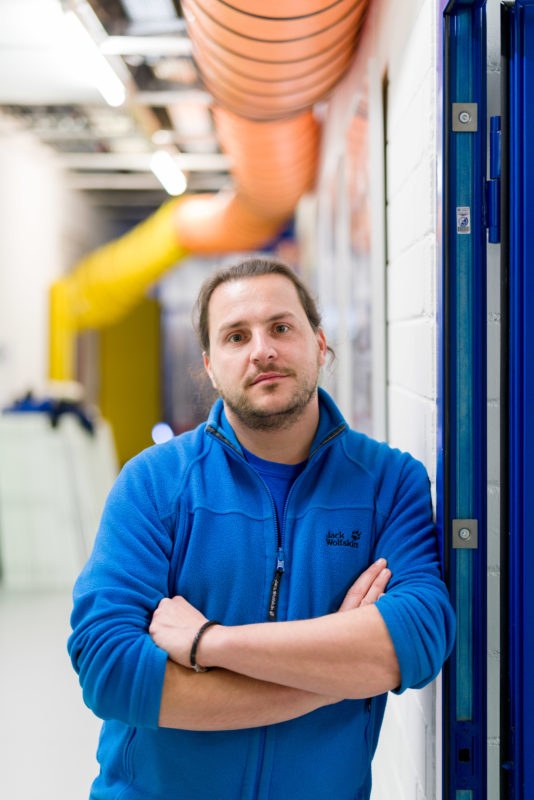 Science photography at a physics institute of the University of Stuttgart: A portrait of one of the researchers. He casually leans against an open door in the corridor. Above it large ventilation pipes.