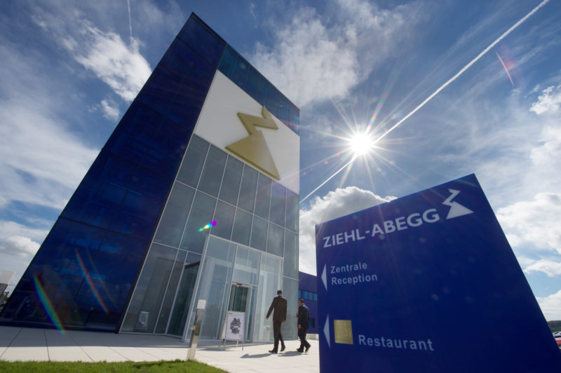 Architekturfotografie: Moderne Firmenzentrale der Firma Ziehl-Abegg von außen. Im Vordergrund sieht man das blaue Firmenschild gegen den blauen leicht bewölkten Himmel mit der strahlenden Sonne, das auf den Eingang des links daneben aufragenden Hauptebäudes weist.