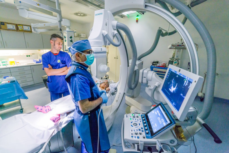 Medizinfotografie: Zwei Ärzte bereiten den Herzkathetereingriff an einem kleinen Mädchen vor, das narkotisiert auf dem Untersuchungstisch liegt. Es ist umgeben von den in alle Richtungen drehbaren Armen der Röntgengeräte. Im Vordergrund sieht man das Ultraschallbild des schlagenden Herzens.