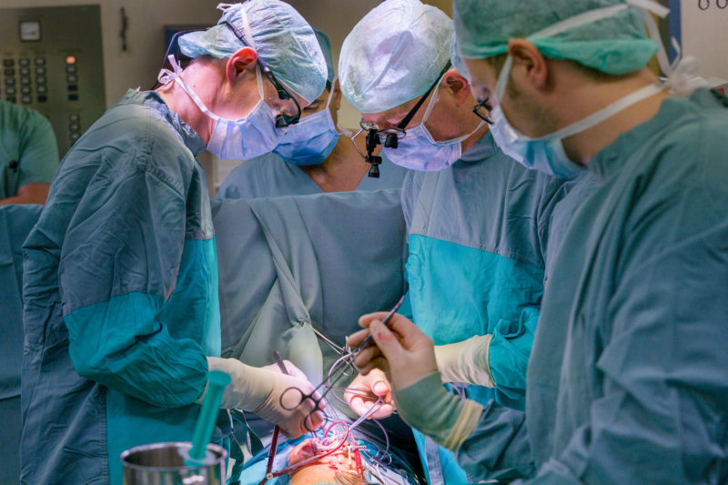 Medizinfotografie: Hochkonzentrierte Ärzte während einer Operation am offenen Herzen eines Kindes. Die chirurgischen Instrumente, klemmen und Schläuche beherrschen das rötlich scheinende Operationsfeld, während das Grün der sterilen Kleidung im Kontrast dazu steht.