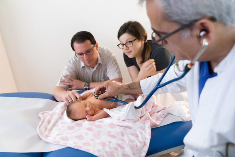 Medizinfotografie: In der kinderkardiologischen Ambulanz der Universitätsklinik Homburg (Saar) untersucht ein Arzt ein Baby. Die besorgten Eltern schauen im Hintergrund zu.